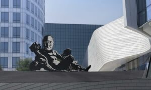 Photo of figural sculpture on cement platform with glass-facade office buildings and the light-colored OCMA building