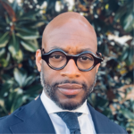 Headshot of J. Kameron Carter in front of foliage, wearing thick-rimmed eyeglasses, jacket and tie