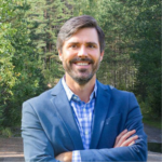 Headshot of James Nisbet in forest setting, arms crossed, wearing checked shirt and blue blazer