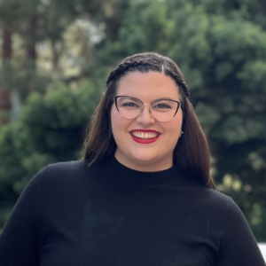 Engler headshot with brown hair and crown braid, eyeglasses, black top, and green foliage background